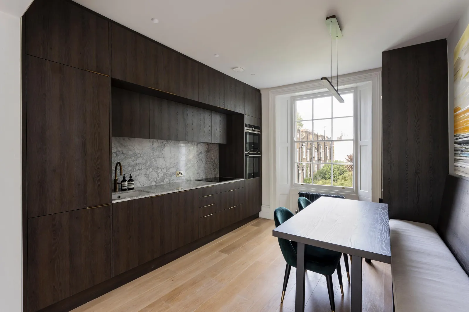 Full-height dark oak kitchen with a grey marble splashback, and a dining table with a built-in banquette by a sash window