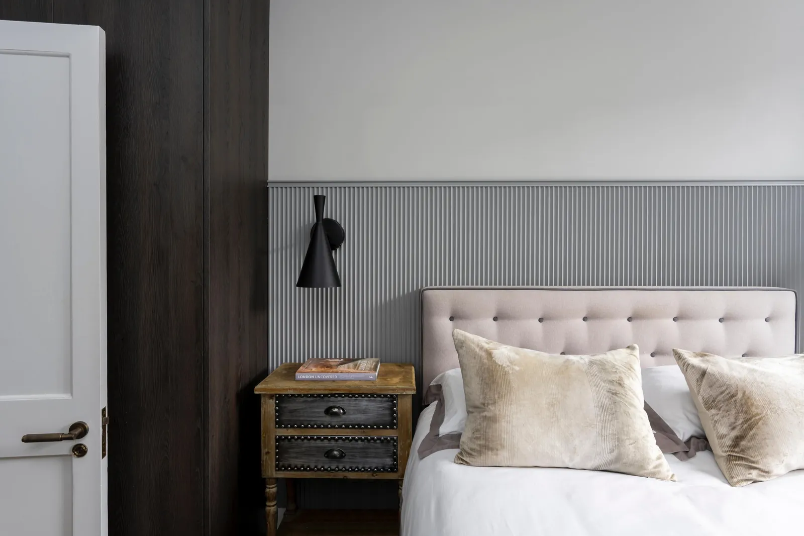 Master bedroom with grey fluted wall panelling, an upholstered headboard, a black wall light and dark oak wardrobes