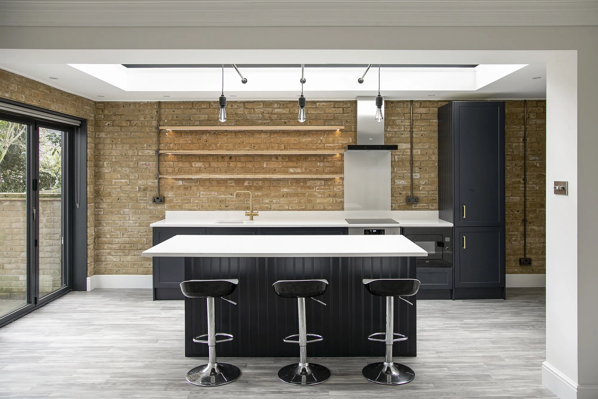 New kitchen in a Fulham rear extension: navy island with a breakfast bar and three stools, exposed London stock brick, oak shelves and a lantern rooflight above