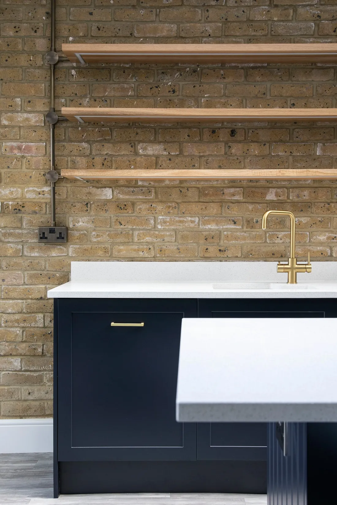 Oak shelves on exposed London stock brick above navy shaker units and a brass tap