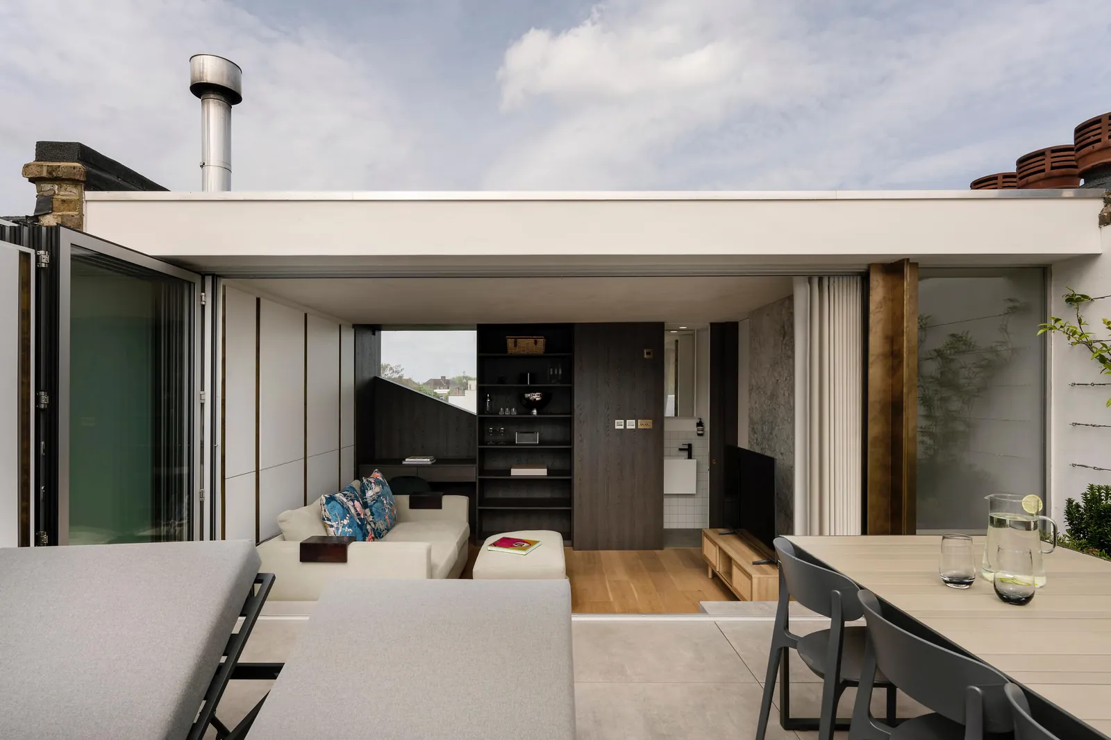 Loft living room seen from the roof terrace through open bifold doors, with dark oak shelving and a guest bathroom beyond